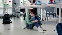 Depressed lonely boy sitting under desk in classroom with classmates playing on background